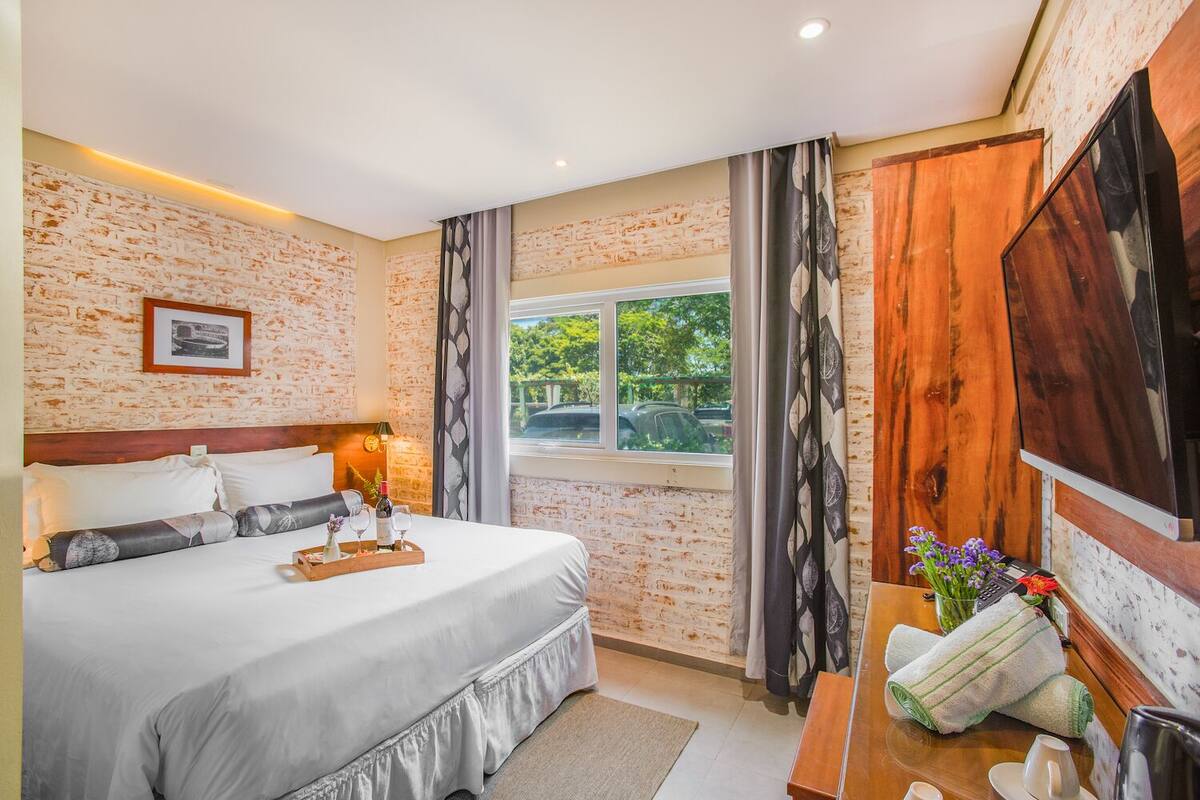 Habitación de hotel con pared de ladrillo, una cama grande con cabecera de madera, un escritorio con computadora y una ventana con vista a follaje.