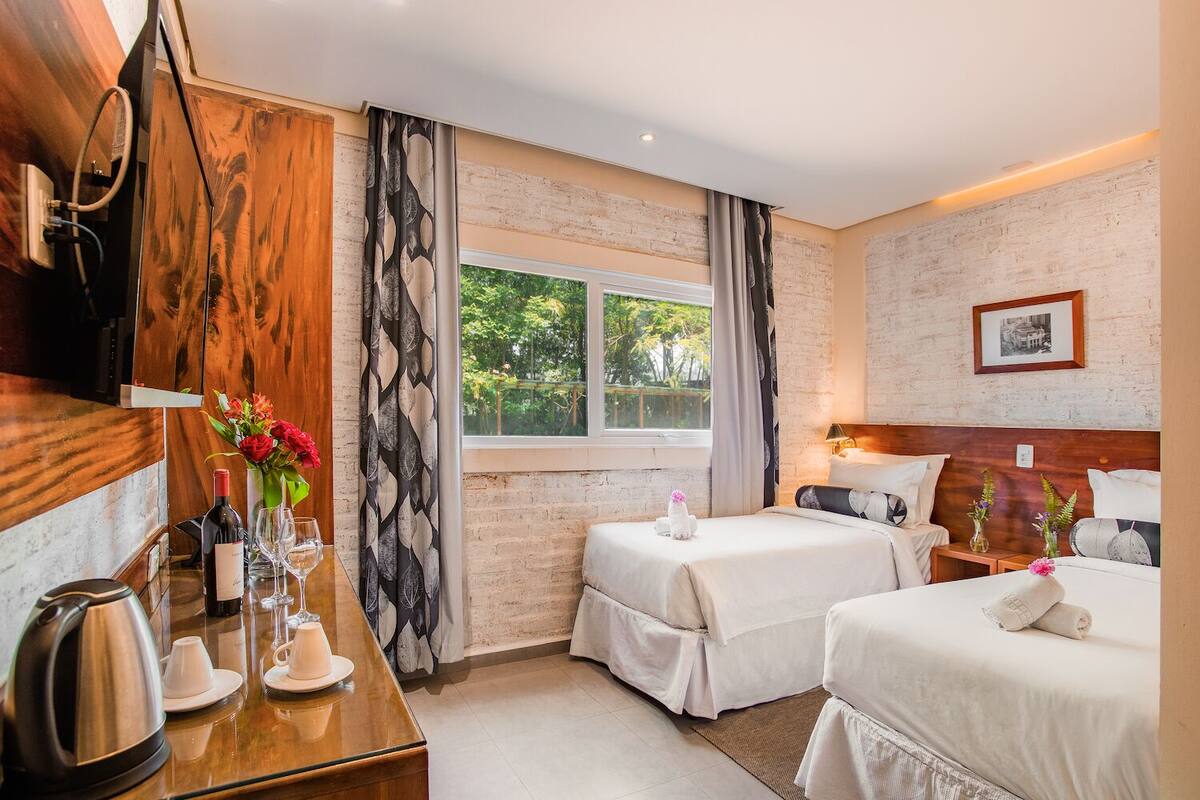 Una habitación de hotel con dos camas, un escritorio de madera con un hervidor, una mesita con un florero y una ventana con vistas a la vegetación.