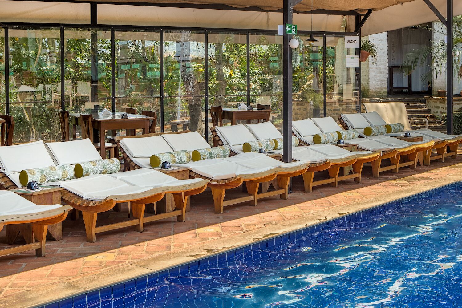 Área à beira da piscina com espreguiçadeiras e mesas, uma piscina revestida em azulejos e vista para o verde através de grandes janelas.