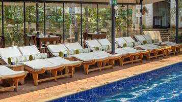 Área à beira da piscina com espreguiçadeiras e mesas, uma piscina revestida em azulejos e vista para o verde através de grandes janelas.