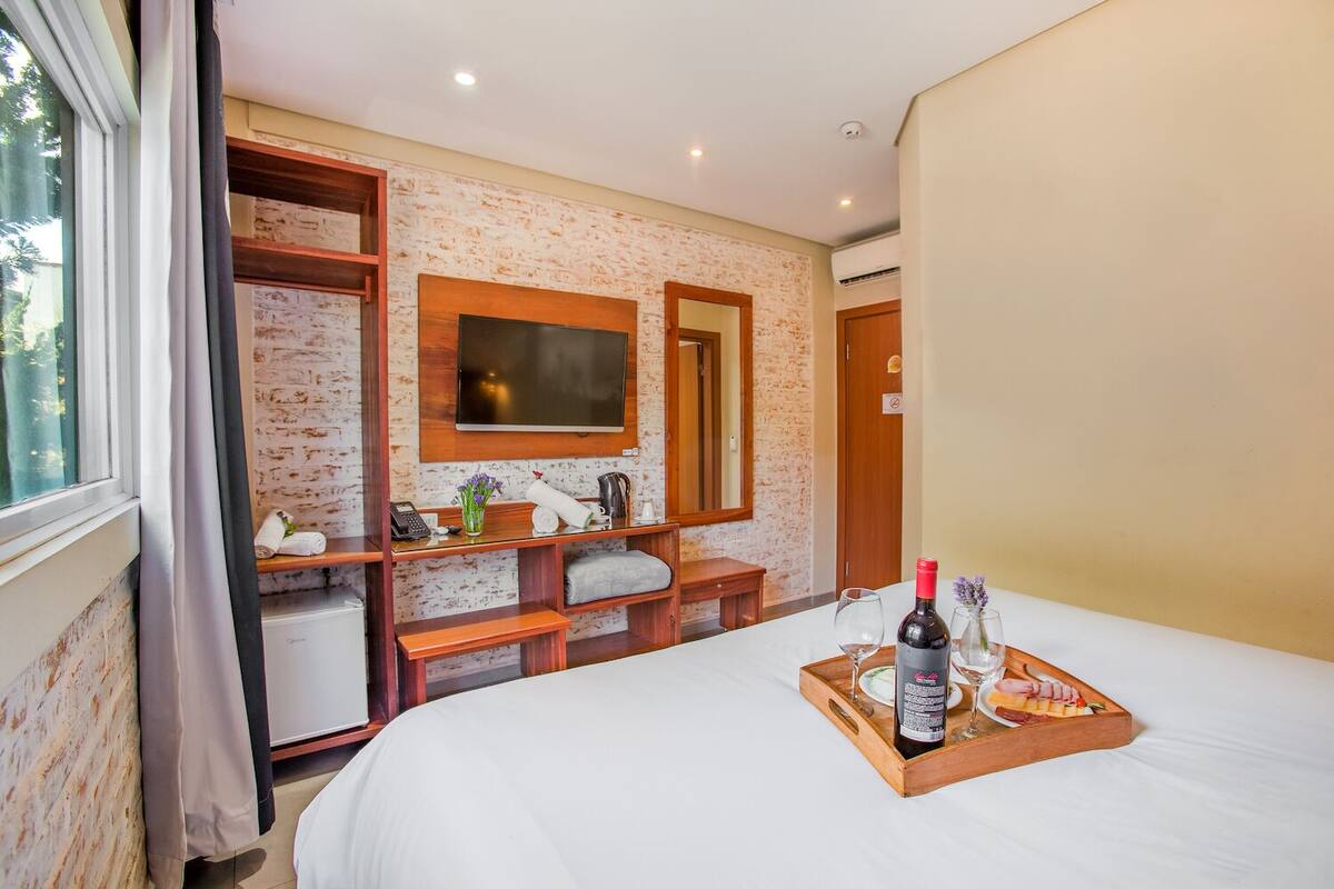 Habitación de hotel con cama, mesita de madera con bandeja de comida y bebidas, televisor montado en una pared de ladrillo, armario de madera y ventana con cortinas.