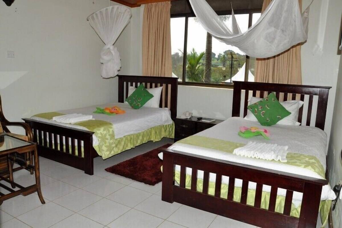 Quarto com duas camas de madeira, cada uma com travesseiro verde e manta branca. Há uma pequena mesa e uma cadeira no canto.