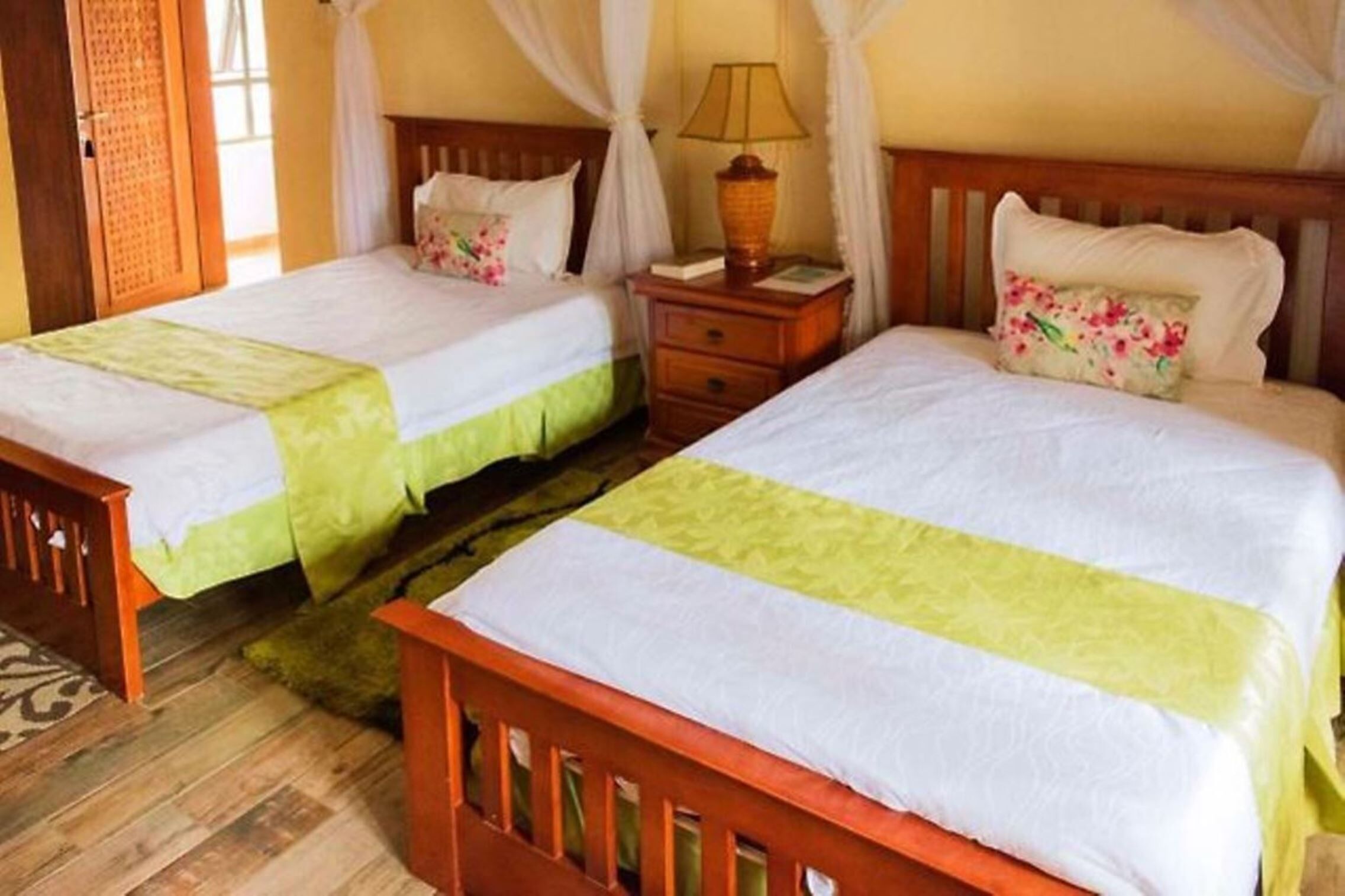 A room with two wooden beds, each with a green bedspread and white pillows. There is a nightstand with a lamp between the beds.