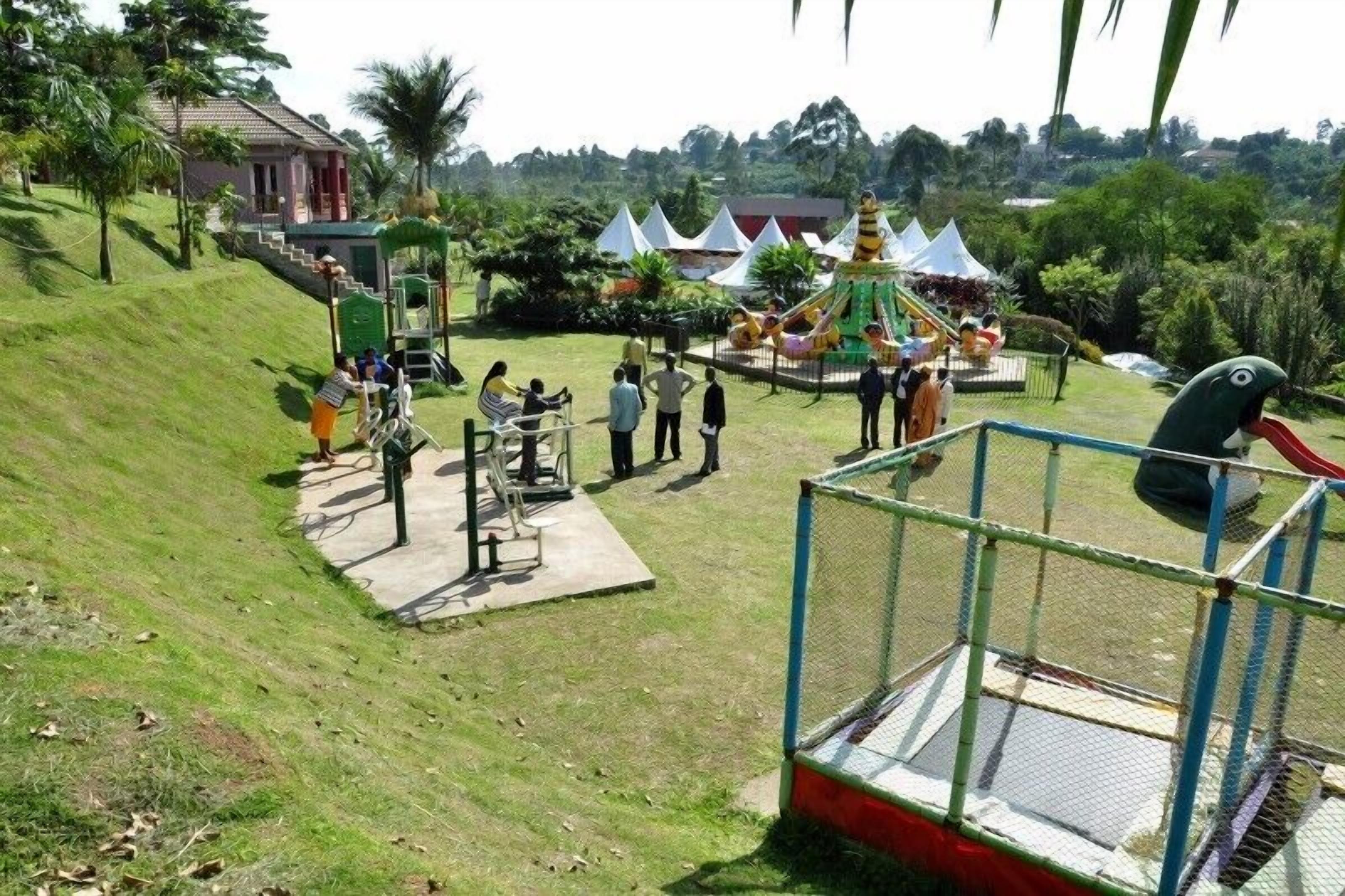 A playground with slides and climbing structures, surrounded by greenery and a few buildings in the background.