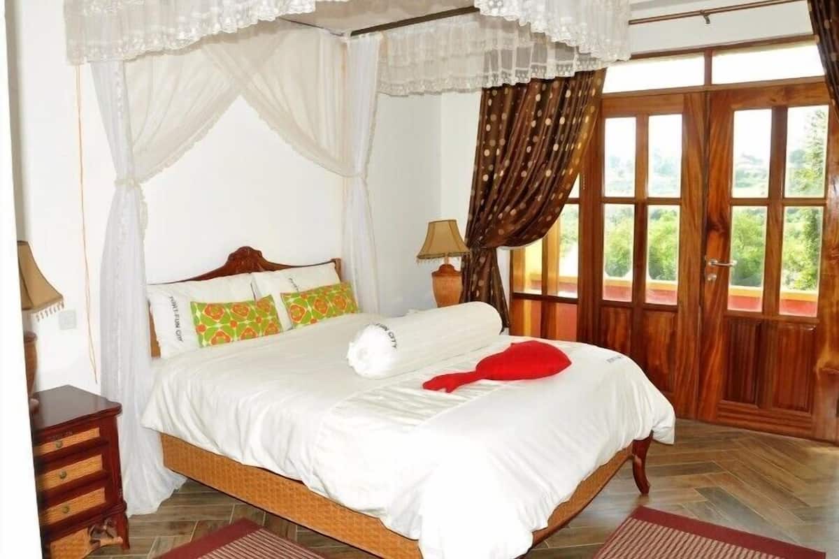 Quarto com cama de dossel, piso de madeira, mesa de cabeceira, abajur e vista para o verde pela janela.