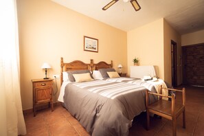 Une chambre à coucher avec un grand lit, deux tables de chevet, une chaise en bois, un ventilateur de plafond et une porte.