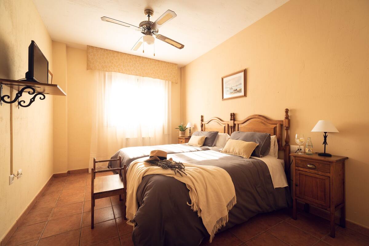 Une chambre à coucher comprenant un lit, des tables de chevet, un ventilateur de plafond, une télévision et une fenêtre avec des rideaux.