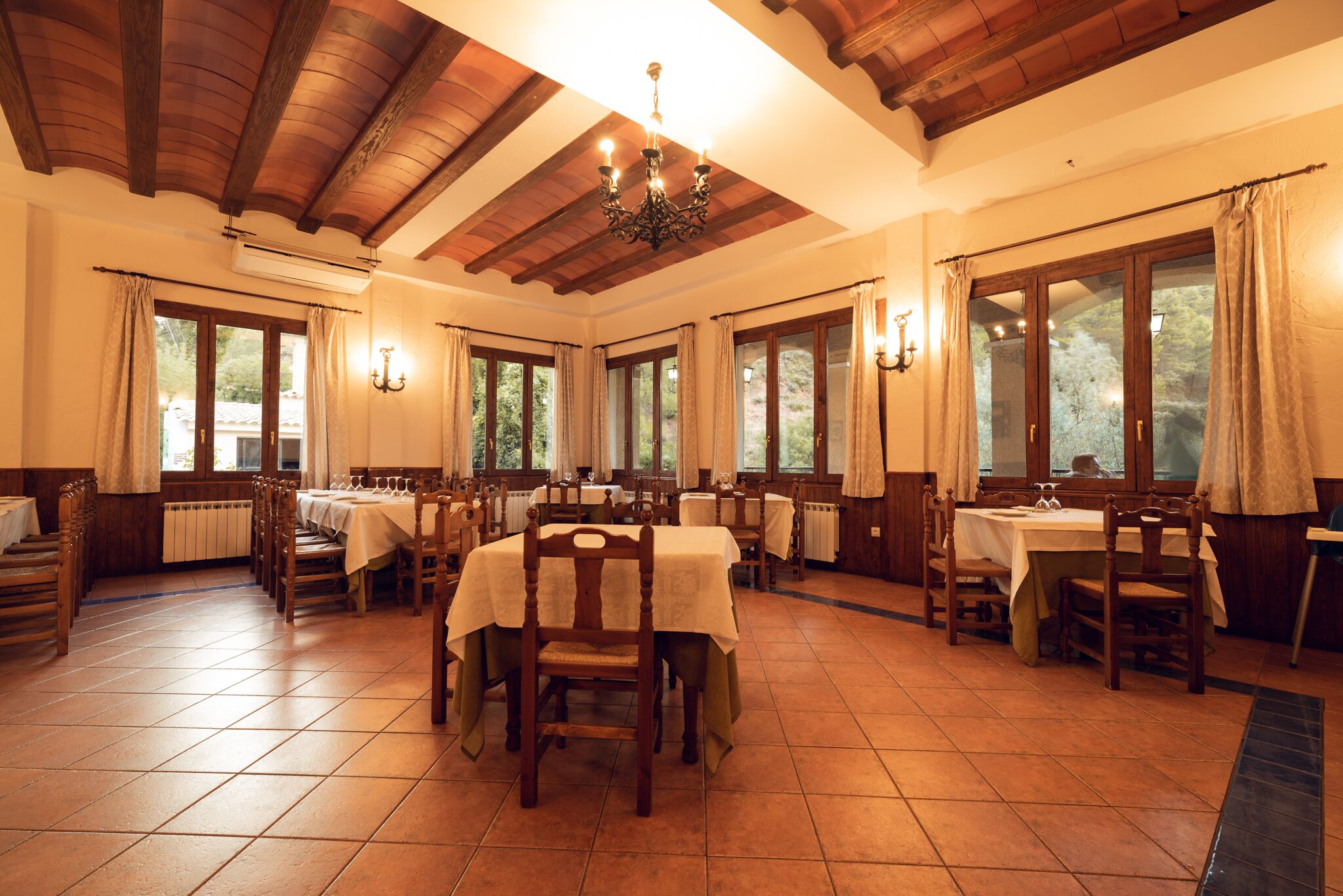 Un comedor con mesas y sillas de madera, ventanas amplias y un candelabro.