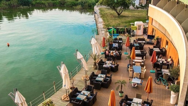 Un espacio al aire libre junto al río con mesas, sillas, sombrillas y plantas en macetas.