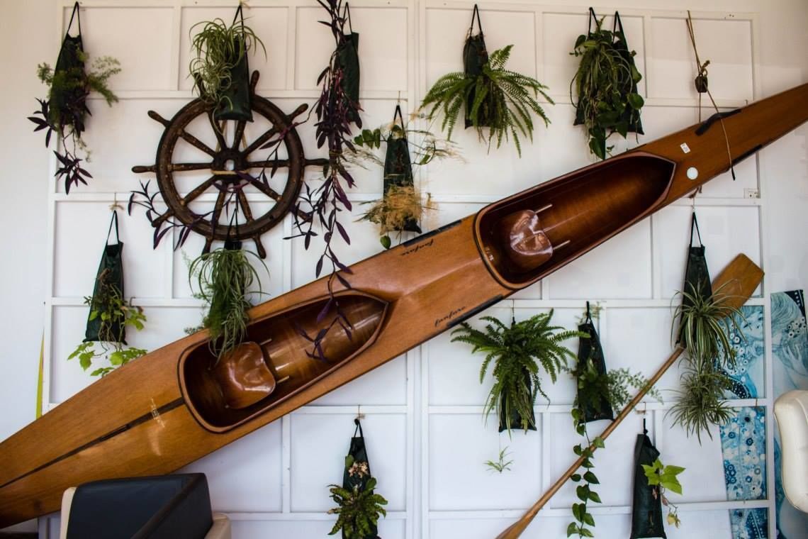 A wooden boat-shaped planter with a steering wheel, surrounded by hanging plants on a white wall.
