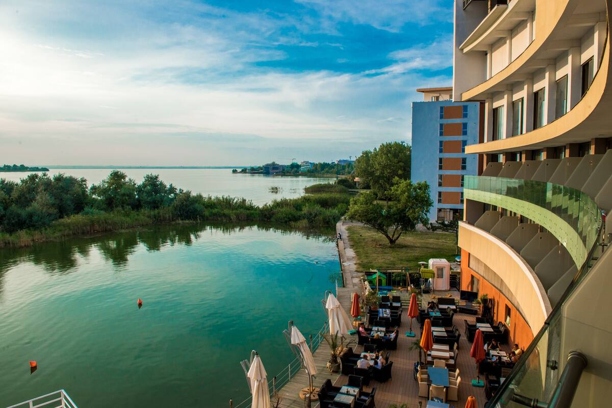 A waterfront hotel with outdoor seating overlooking a calm river, surrounded by greenery and modern architecture.