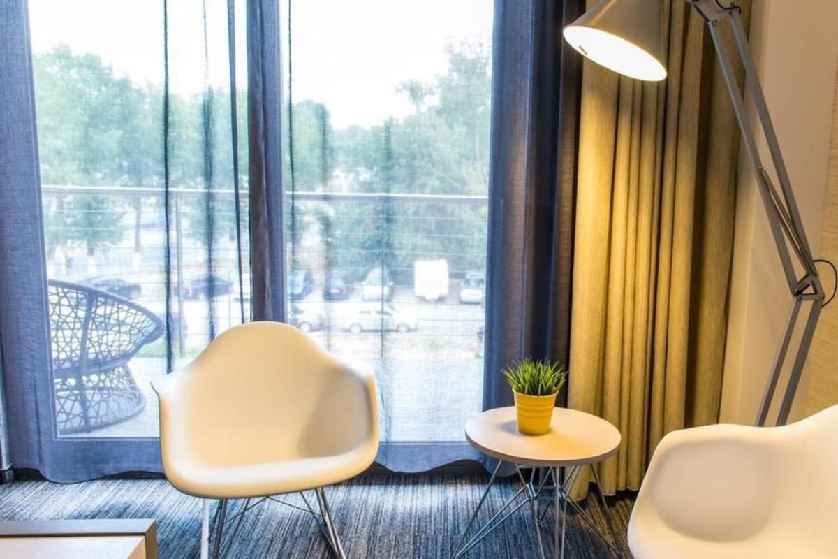 A hotel room with two white chairs, a small round table with a potted plant, a floor lamp, and a view of a parking lot through large windows.