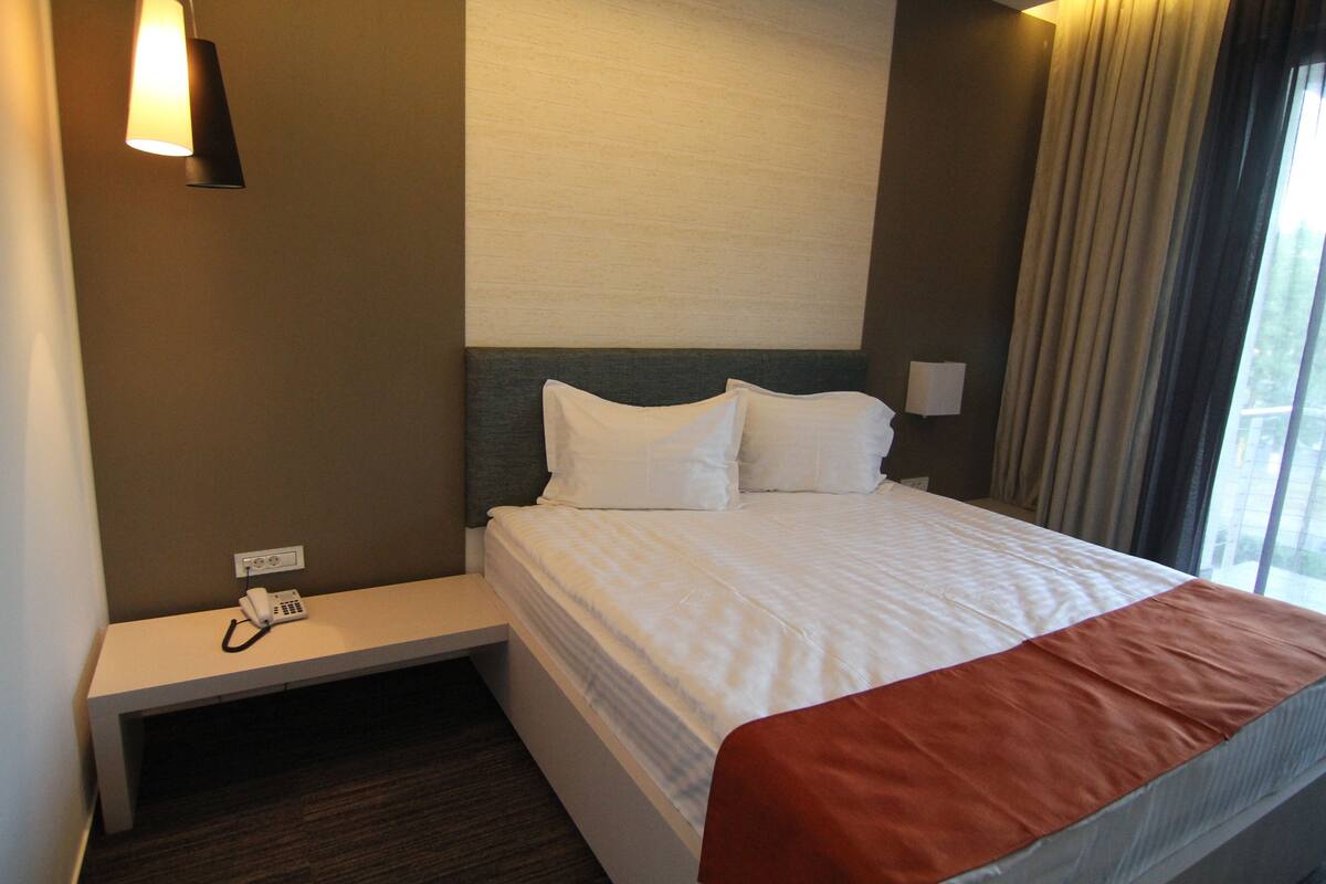 A hotel room with a bed, bedside table, lamp, and a window with curtains.