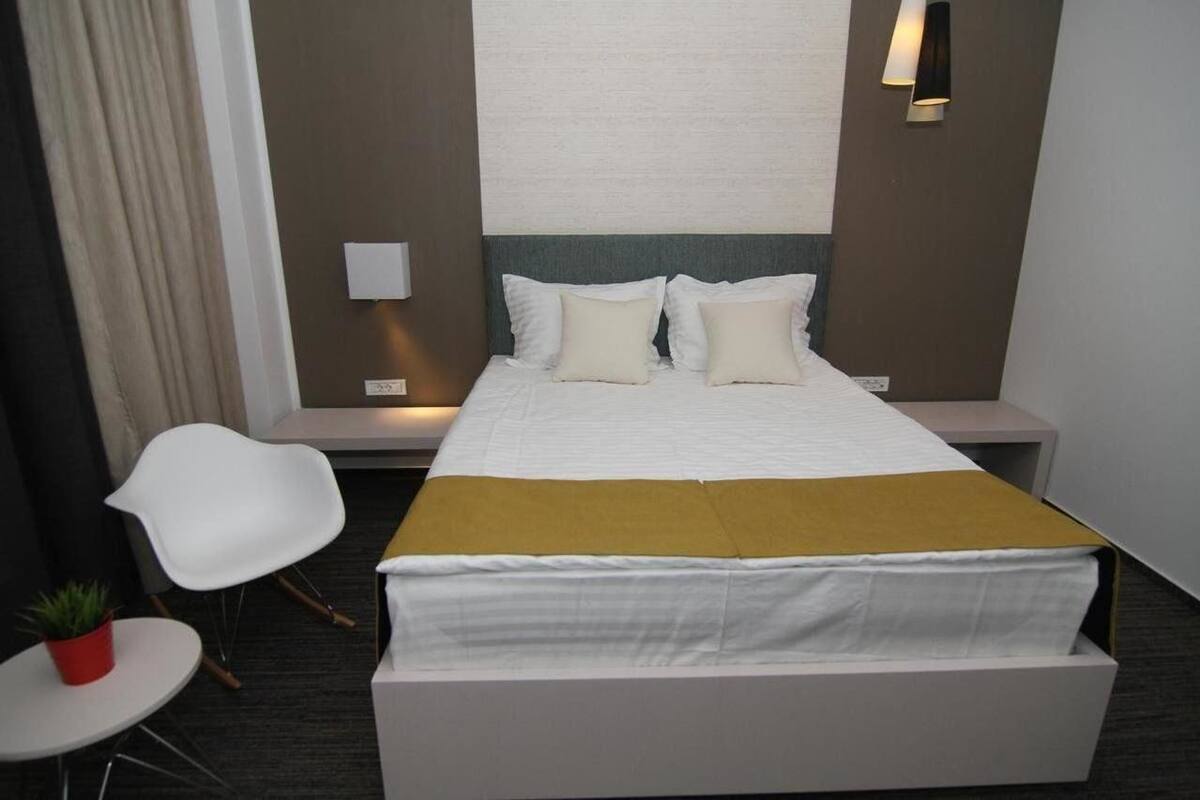 A hotel room with a bed, a white chair, a small table with a plant, and a wall-mounted light fixture.