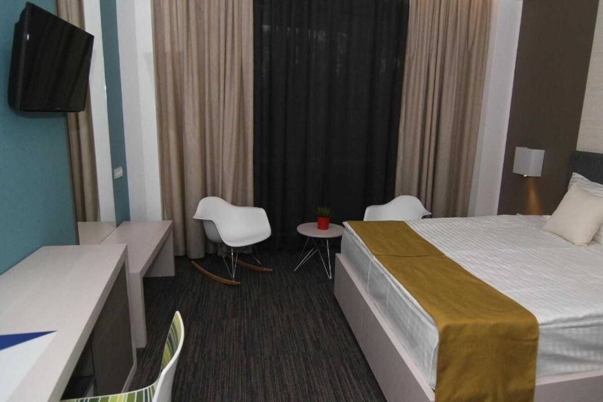 A hotel room with a bed, a desk, a chair, a small table with a plant, and a TV mounted on the wall.