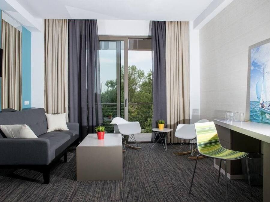 A modern hotel room with a grey sofa, a small coffee table, a TV, a balcony with curtains, and a painting of a sailboat.