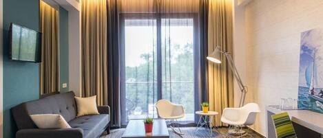 Habitación de hotel moderna con sofá, una mesita con planta, escritorio con silla y una ventana grande con cortinas.