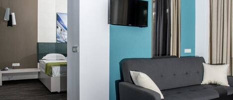 A modern hotel room with a grey sofa, a flat-screen TV, a bed with a green blanket, and a window with curtains.