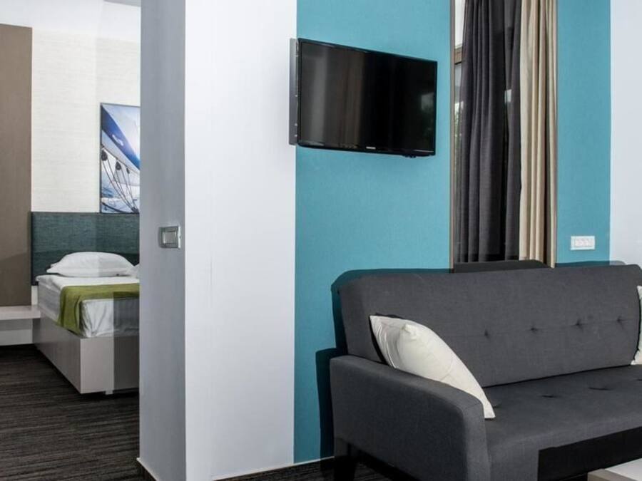 Ein modernes Hotelzimmer mit grauem Sofa, einem Flachbildfernseher, einem Bett mit grüner Decke und einem Fenster mit Vorhängen.