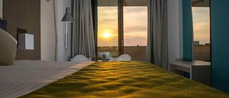 A hotel room with a large bed, a desk, and a view of the sunset through the window.