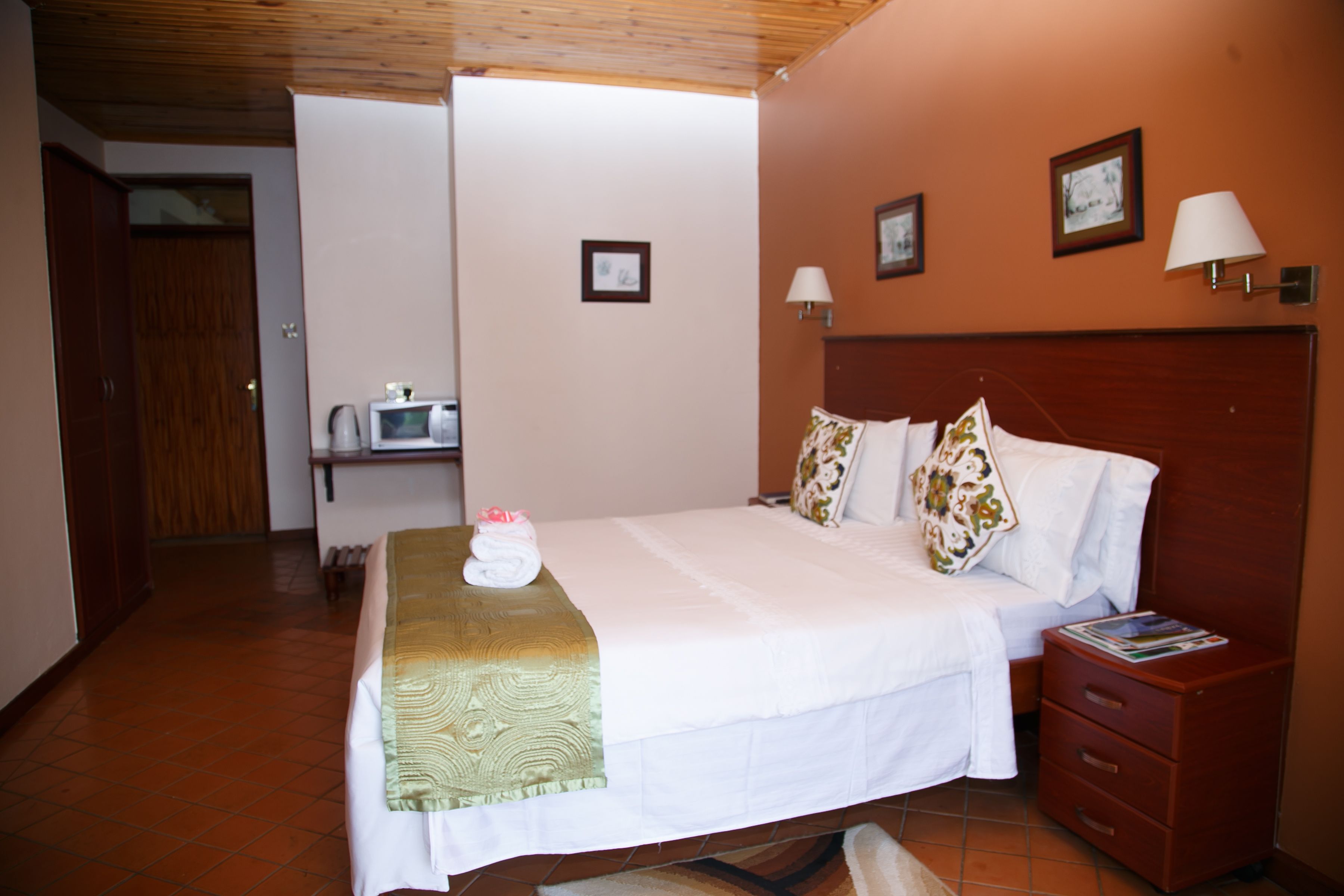 A neatly made bed with a green blanket, a wooden nightstand with a magazine, and a wall-mounted lamp.