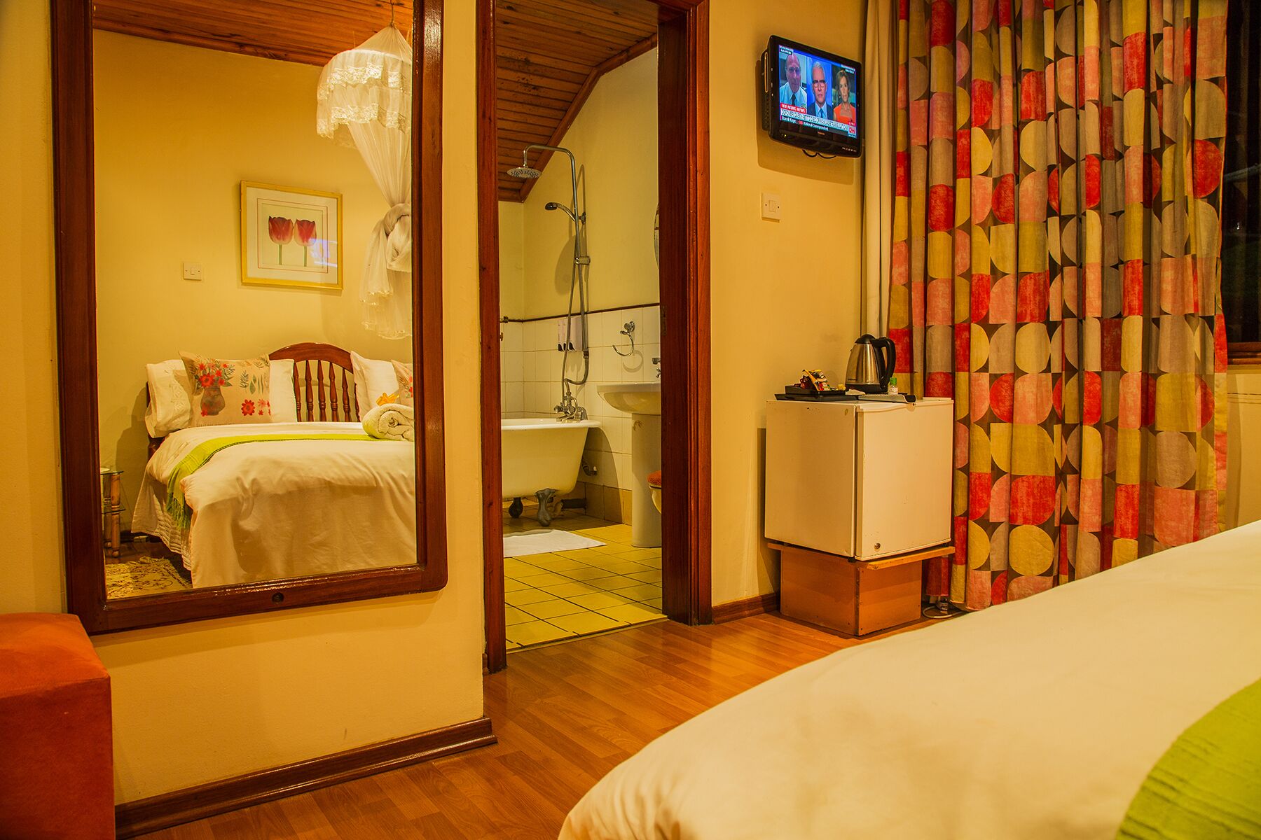 A hotel room with a bed, a small bedside table, a mirror, a television, and a bathroom with a sink and shower.