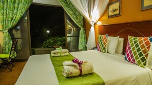 Habitación de hotel con cama, almohadas y colcha verde. Hay una silla y una mesa con una toalla encima.