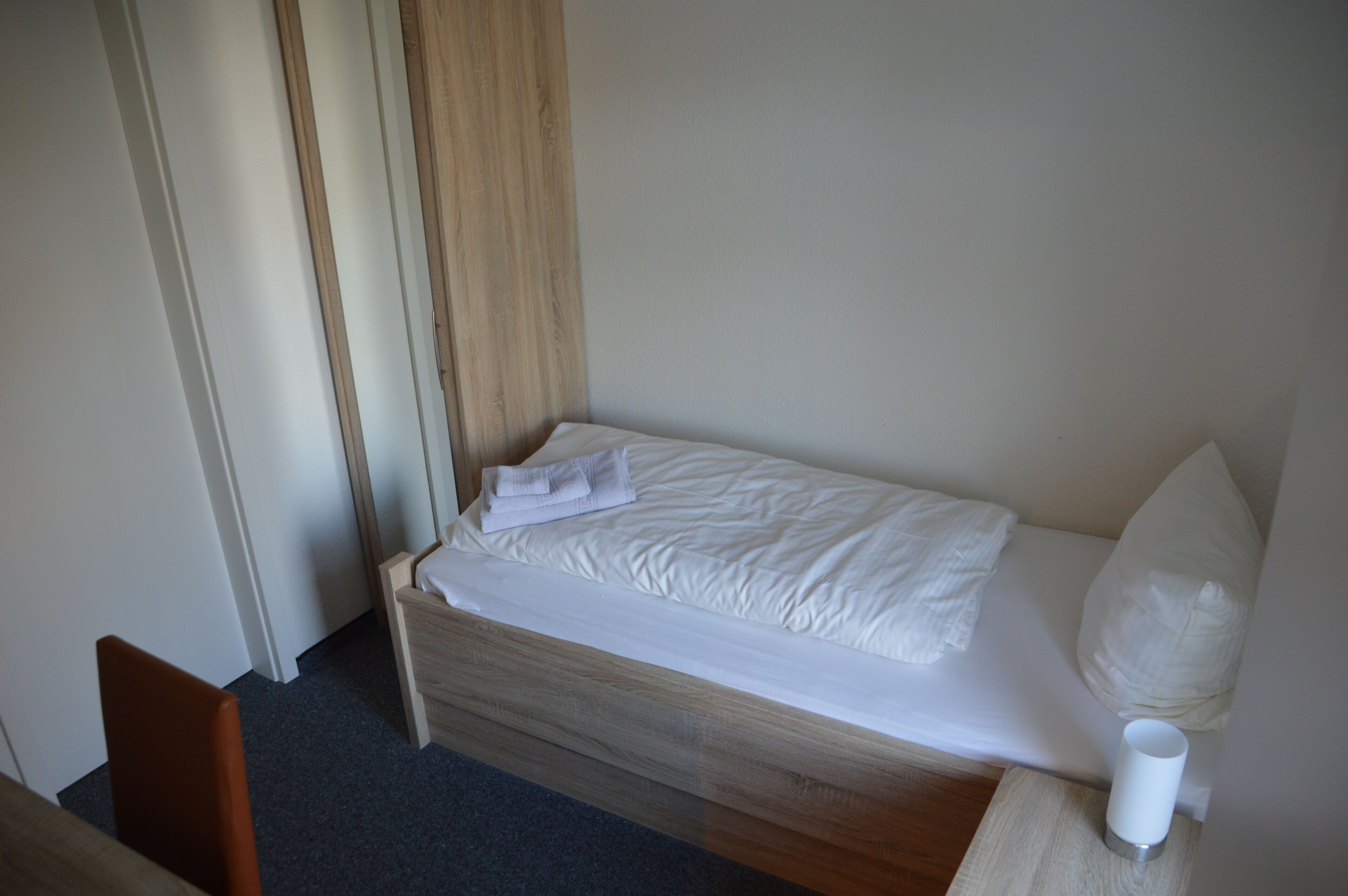 A small, neatly made bed with white linens and pillows, a wooden headboard, and a bedside table with a white object.