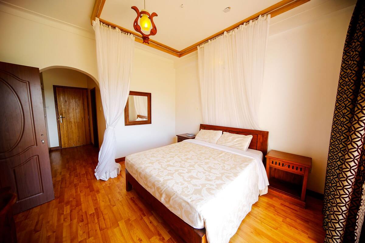 A bedroom with a wooden bed, white curtains, a wooden nightstand, and a mirror on the wall.