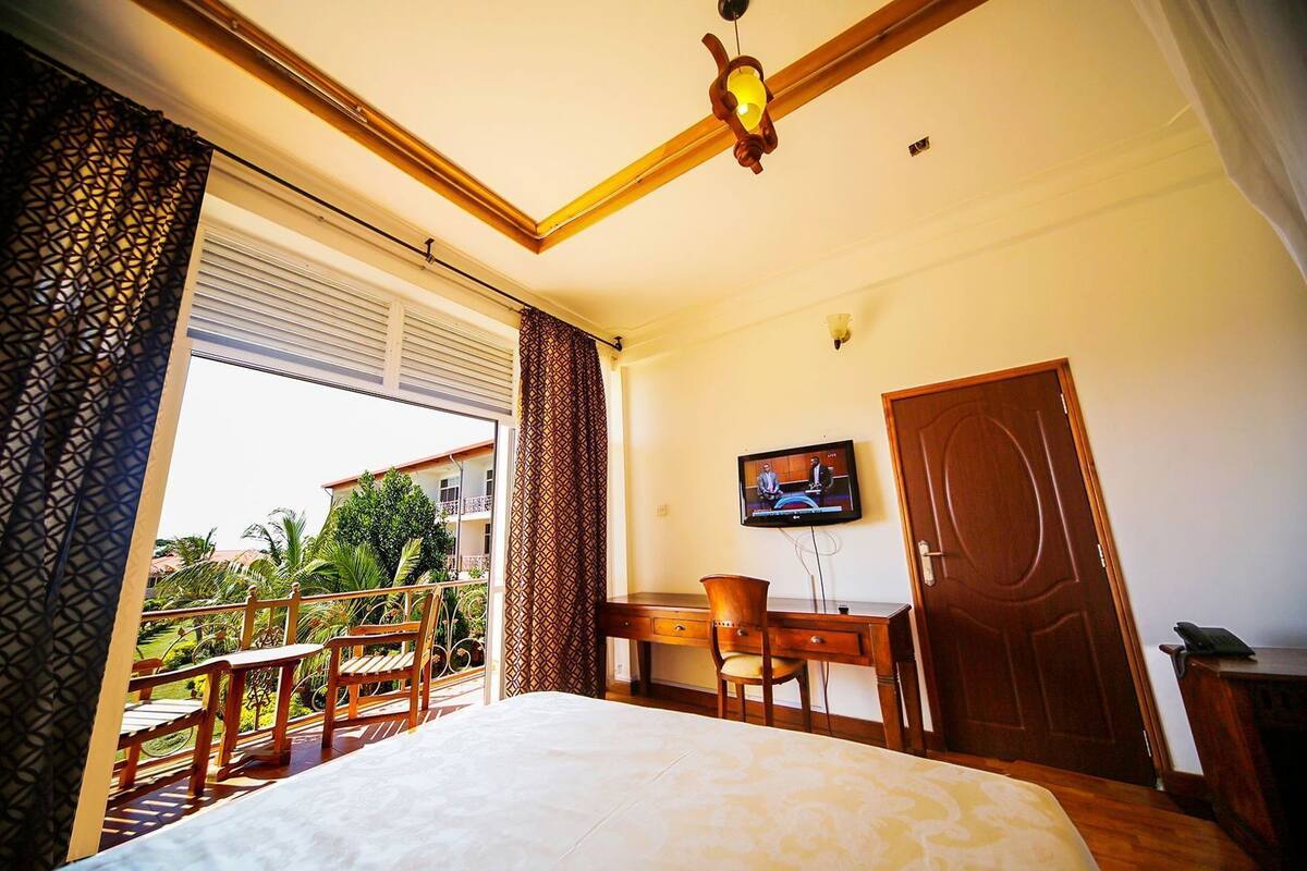 A hotel room with a bed, a desk with a chair, a television, and a balcony with a view of buildings and greenery.