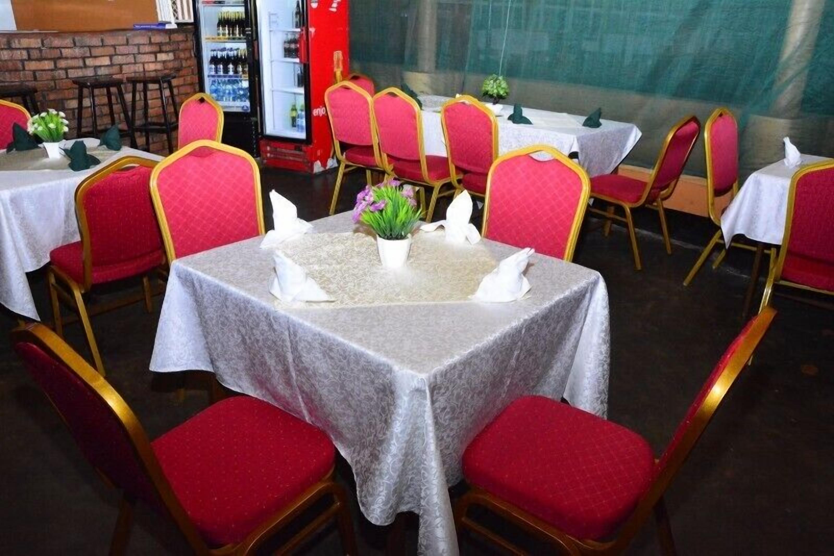 Een restaurant met ronde tafels met witte tafelkleden, rode stoelen met kussens, en een kleine potplant op elke tafel.