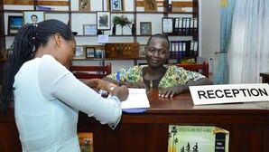 A reception desk with a sign labeled "RECEPTION" and a background of framed pictures and binders. - Mirembe Resort Beach Hotel (Bugala Island)