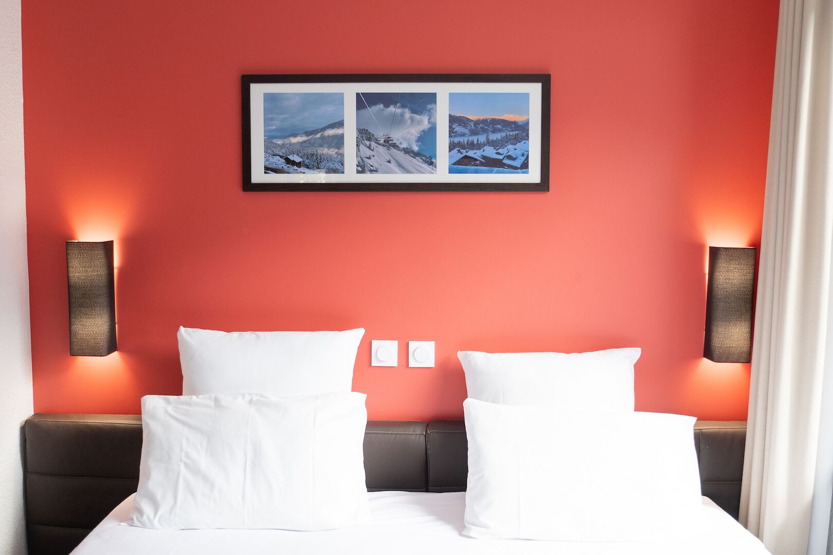 A hotel room with a red wall, a bed with white bedding, two wall-mounted light fixtures, and a framed picture above the bed.