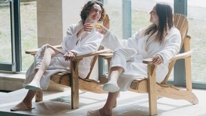 Two people in white robes seated on wooden chairs by a window with a view of a snowy landscape. - Hôtel Le Montana (Courchevel)
