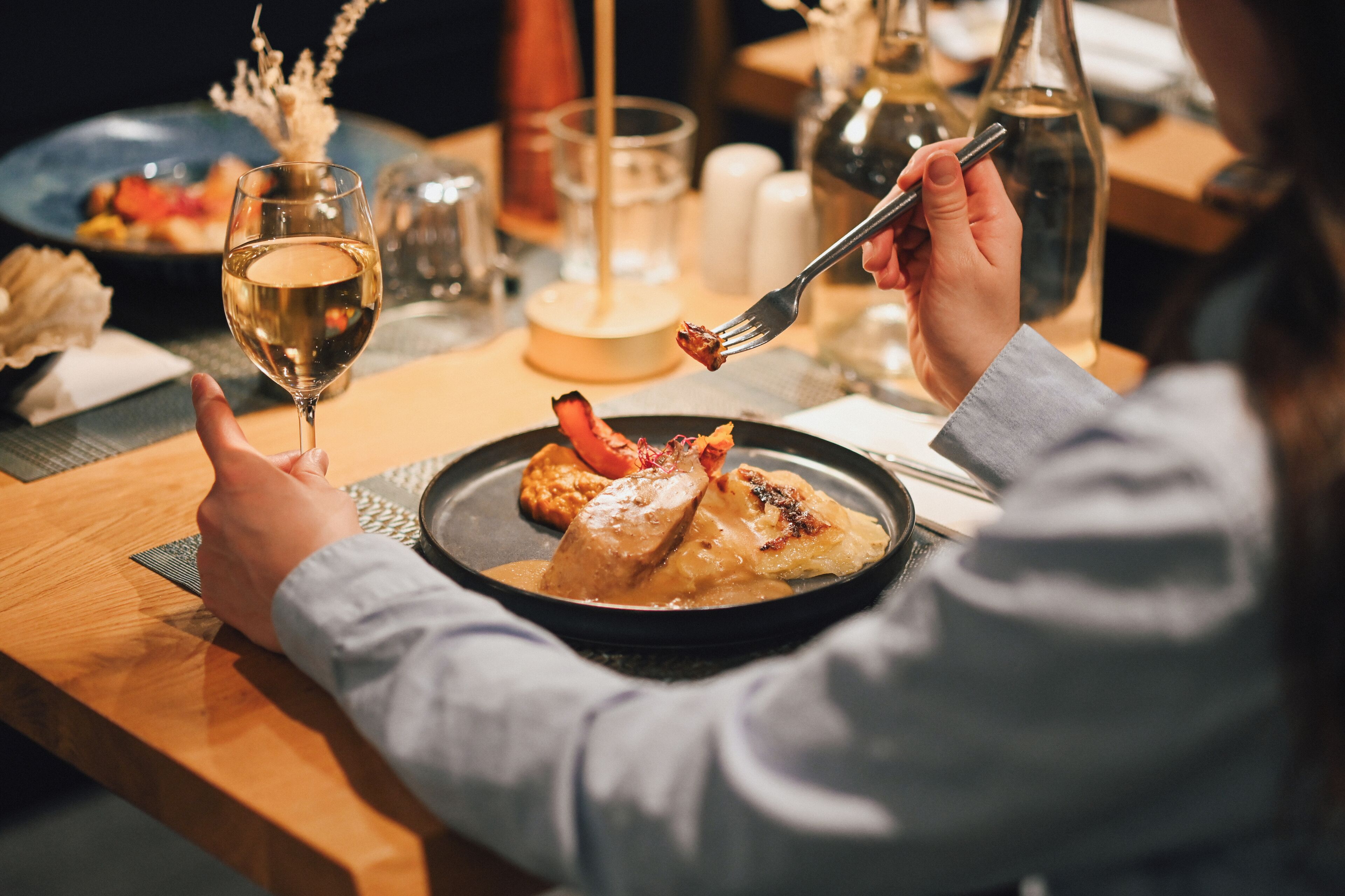 A person dining at a table with a plate of food, a glass of white wine, and a bottle of water.