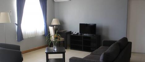 Una sala de estar con sofá, una mesita de noche con lámpara, televisor sobre un mueble bajo y ventana con cortinas.