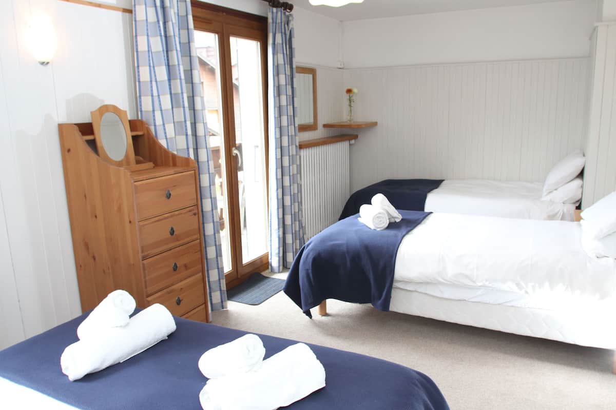 A bedroom with two beds, a wooden dresser with a mirror, a small shelf with a flower, and a door with curtains.