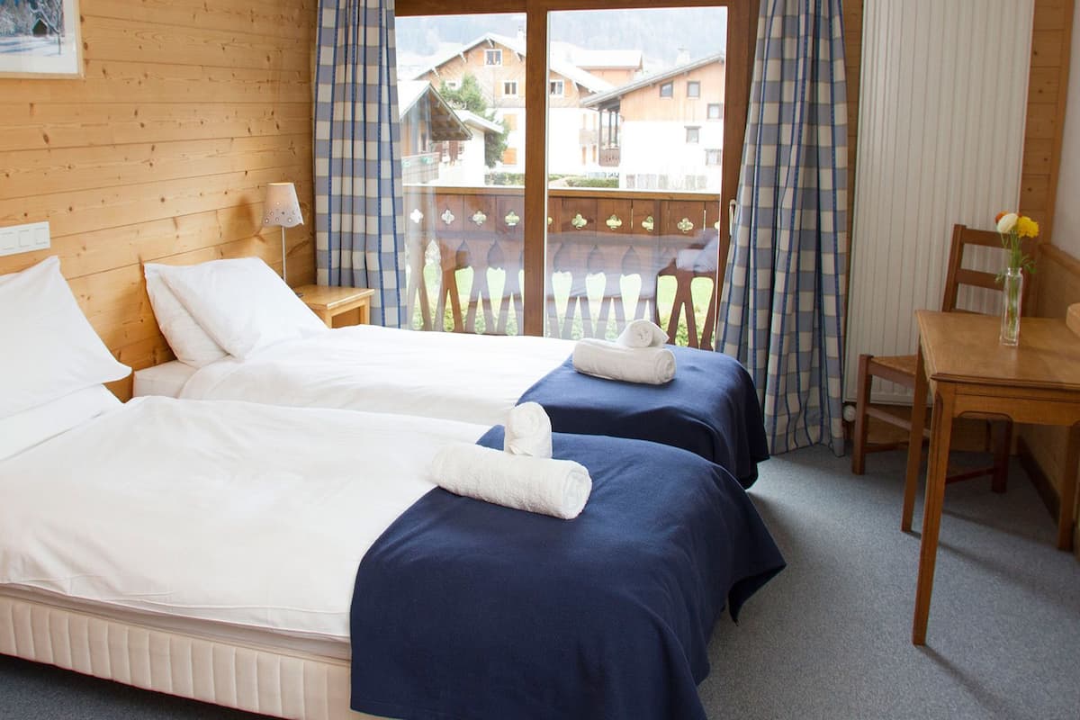 A hotel room with two beds, a wooden headboard, a small table, and a view of the outside balcony.