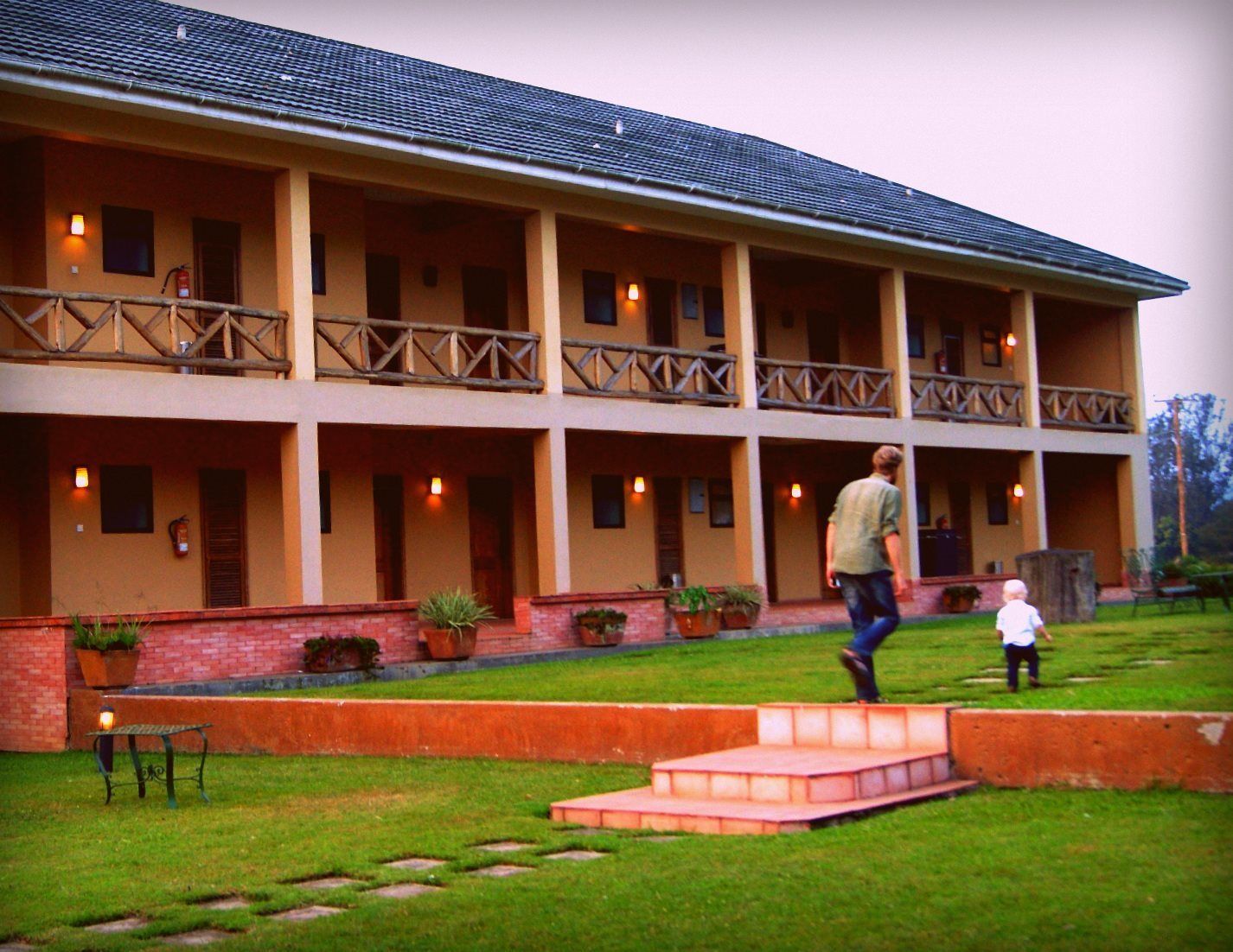 A two-story building with a balcony on each floor, potted plants, and a grassy lawn.