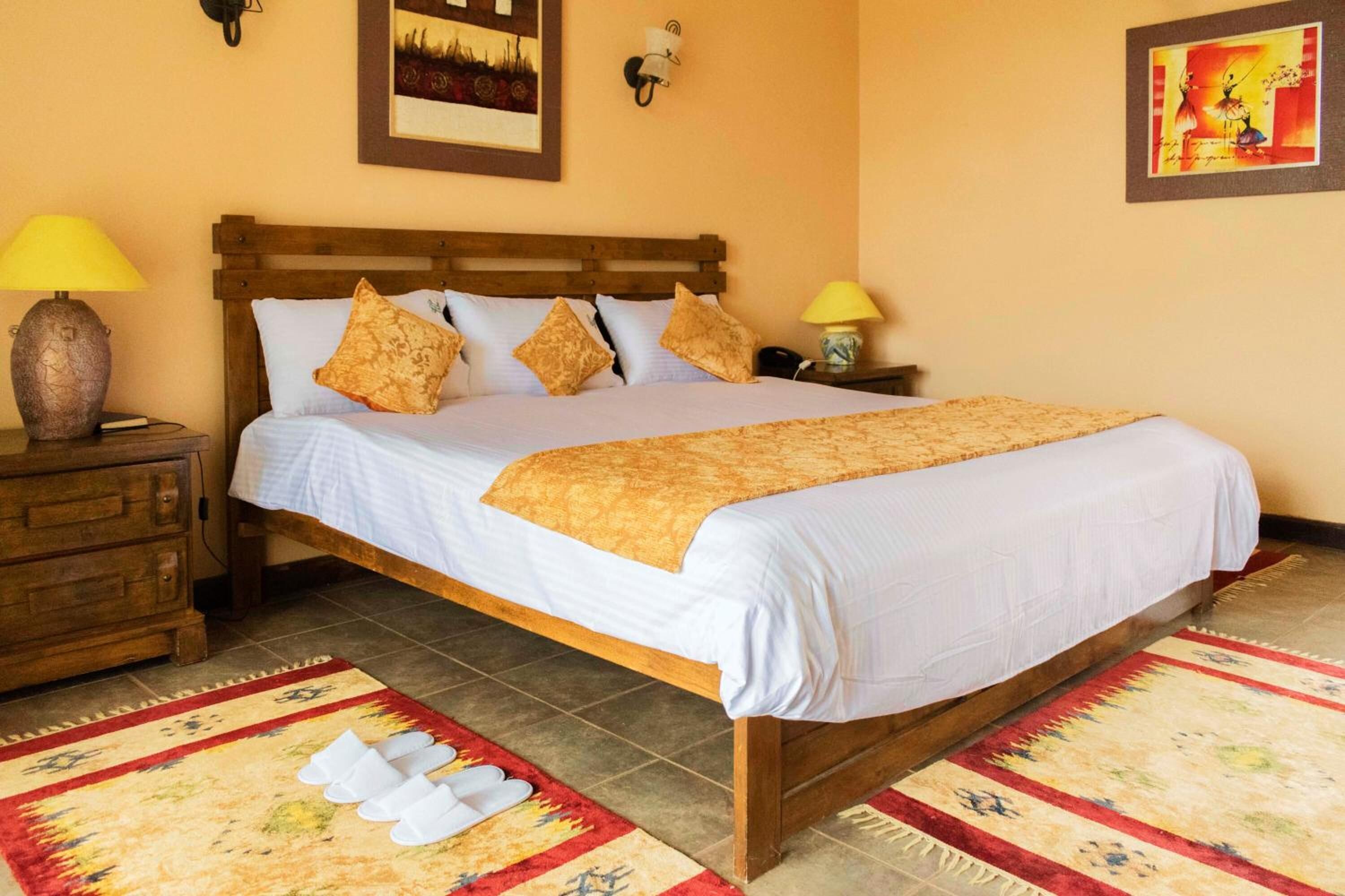 A neatly made bed with a wooden headboard, white bedding, and decorative pillows. There are two bedside lamps, a wooden nightstand with a lamp, and a patterned rug on the floor.