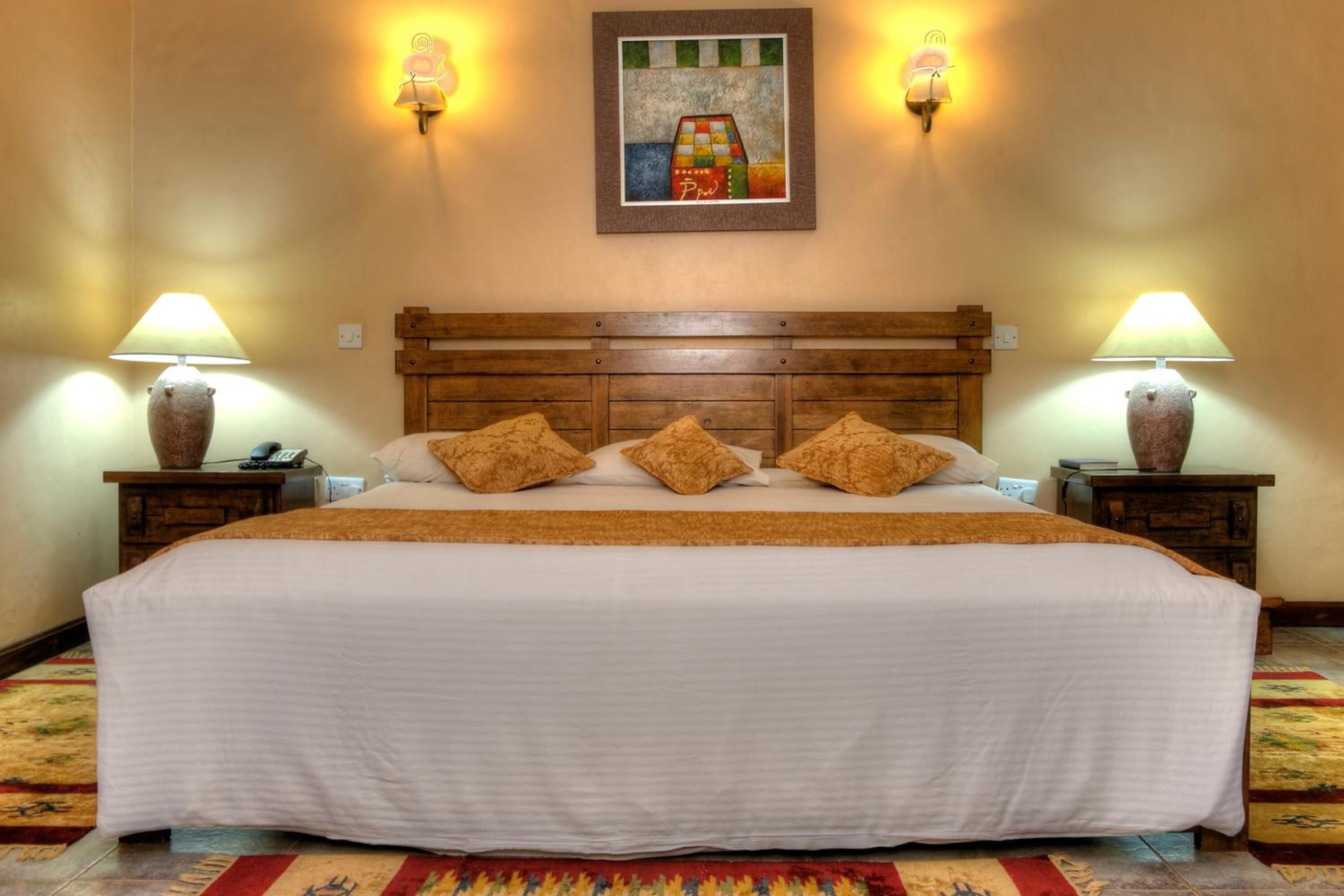 Uma cama grande com cabeceira de madeira, duas mesas de cabeceira com abajures e um tapete colorido.