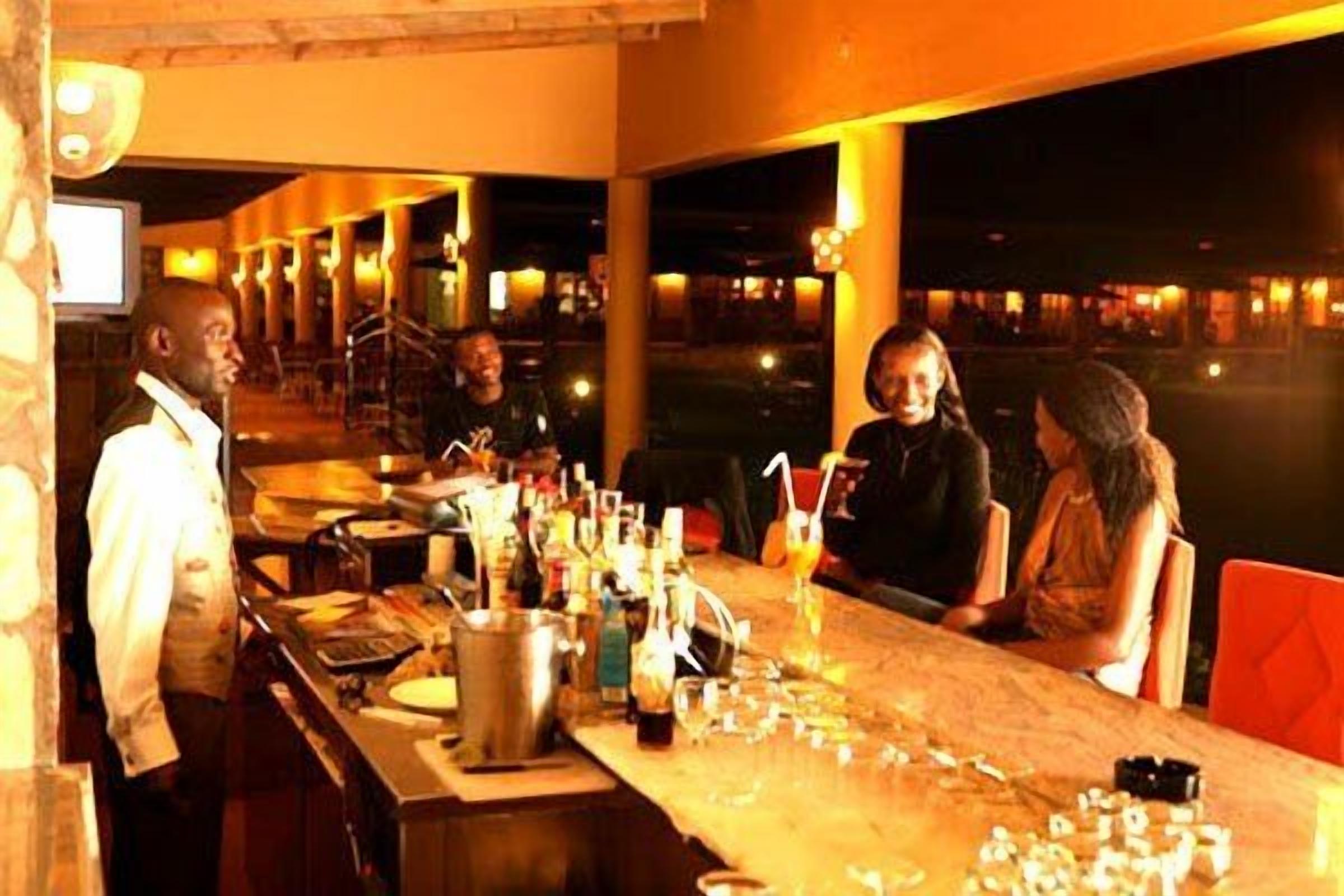 A bar area with a bartender, a counter with various bottles and glasses, and a view of the outside at night.