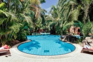 Een rond zwembad omringd door weelderige begroeiing en rotsen, met ligbedden en een jacuzzi.