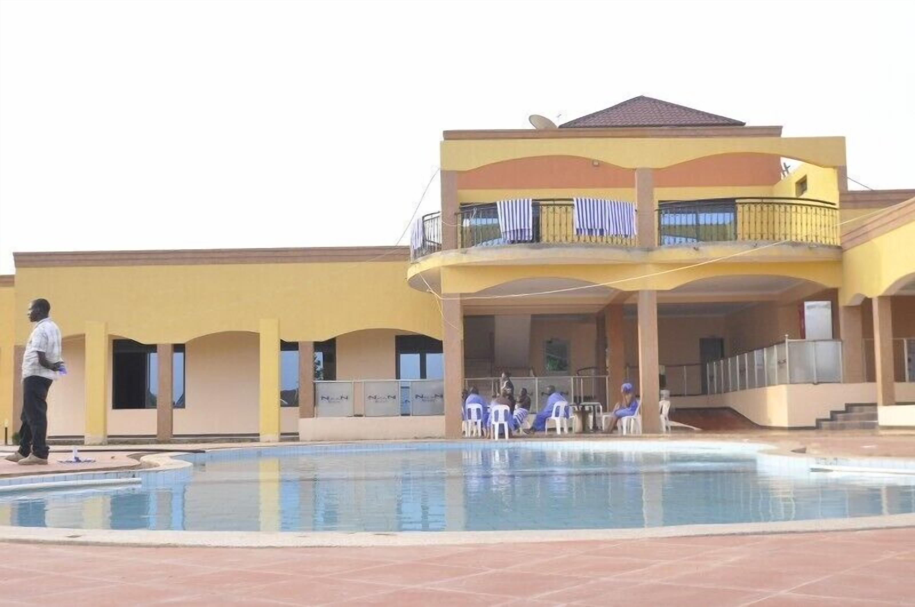 A building with a swimming pool, a covered area with chairs, and a person standing by the pool.