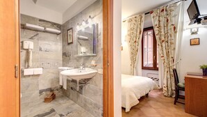 A bathroom with a marble floor, two sinks, a mirror, and a towel rack. The bedroom has a bed, a desk, and a window with curtains. - Hotel Gea di Vulcano (Rome)