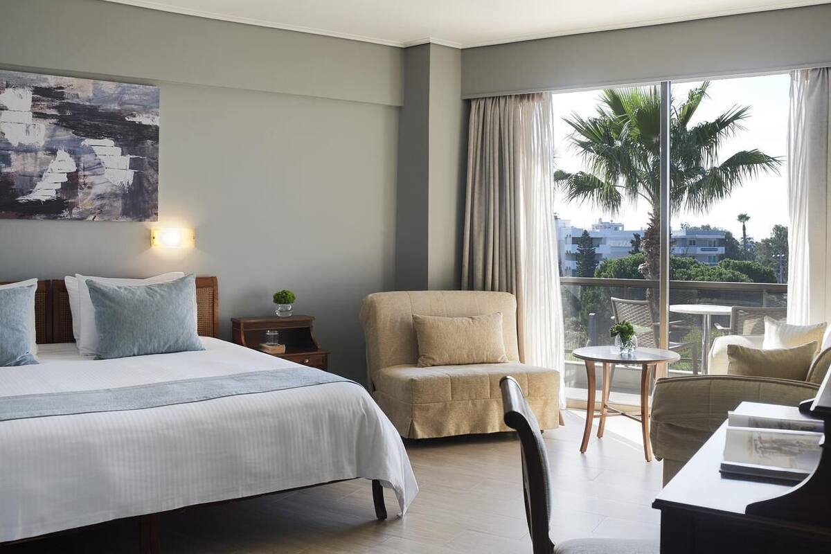 A hotel room with a bed, a chair, a small table, and a view of palm trees and buildings.