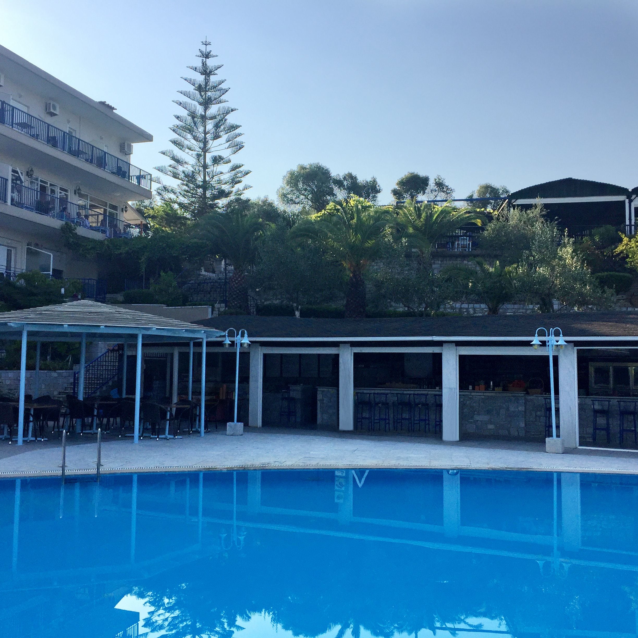 A swimming pool with a covered seating area, a tall tree, and a building in the background.