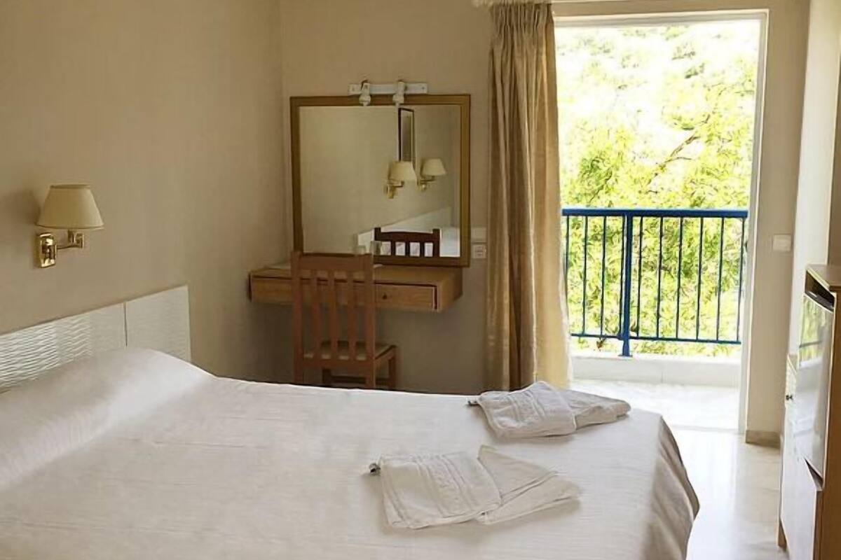 A hotel room with a large bed, a wooden desk with a mirror, a wall-mounted lamp, and a balcony with a view of greenery.