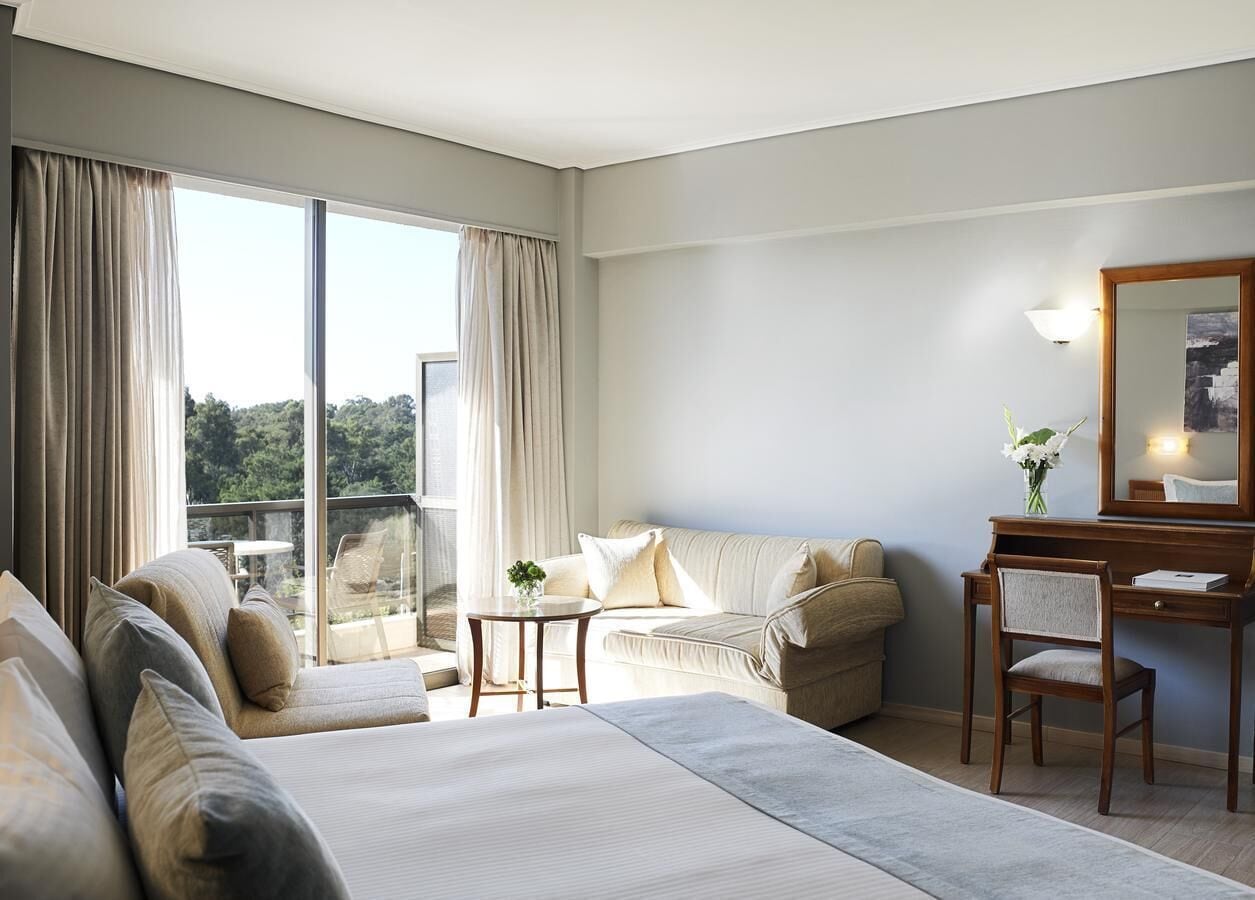 Habitación de hotel con una cama grande, un sofá, un escritorio con silla y un ventanal que ofrece vistas a zonas verdes.