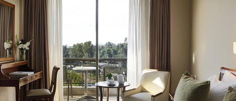 A hotel room with a large bed, a desk with a chair, a small table with a plant, and a view of the outdoors through a sliding door.